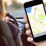female hand holding a phone with interface navigator on a screen on a background woman driving a car